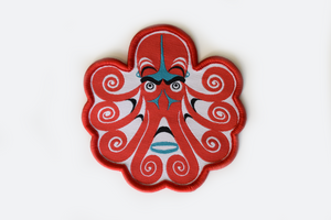 A small patch with a red formline octopus over a white background, it has teal and black accents, and red binding around the edges.