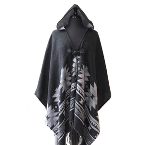 A black hooded shawl draped over a dress form. The shawl features a gray geometric design along the bottom half. It has a toggle button closure, gray lining, and fringe along the bottom hem.